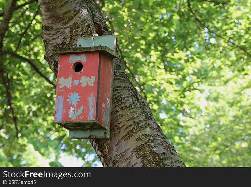 Red birdhouse with painted decorations hangs from tree trunk with green foliage in the background. Red birdhouse with painted decorations hangs from tree trunk with green foliage in the background.