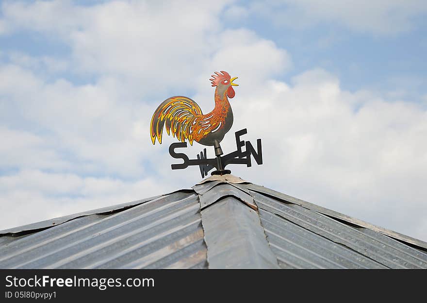 Colorful weather vane with cock figure on the roof over cloudy sky