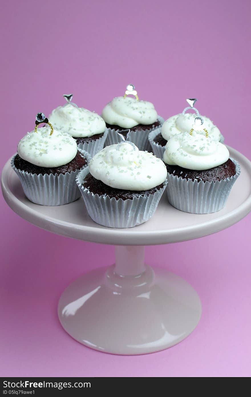 Wedding theme chocolate red valvet cupcakes on pink stand with diamond rings bridal decorations and white frosting against a pink background. Vertical. Wedding theme chocolate red valvet cupcakes on pink stand with diamond rings bridal decorations and white frosting against a pink background. Vertical.