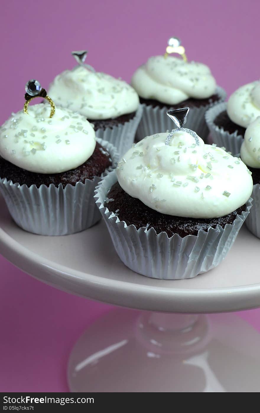 Wedding theme chocolate red valvet cupcakes on pink stand with diamond rings bridal decorations and white frosting against a pink background. Close up. Wedding theme chocolate red valvet cupcakes on pink stand with diamond rings bridal decorations and white frosting against a pink background. Close up.