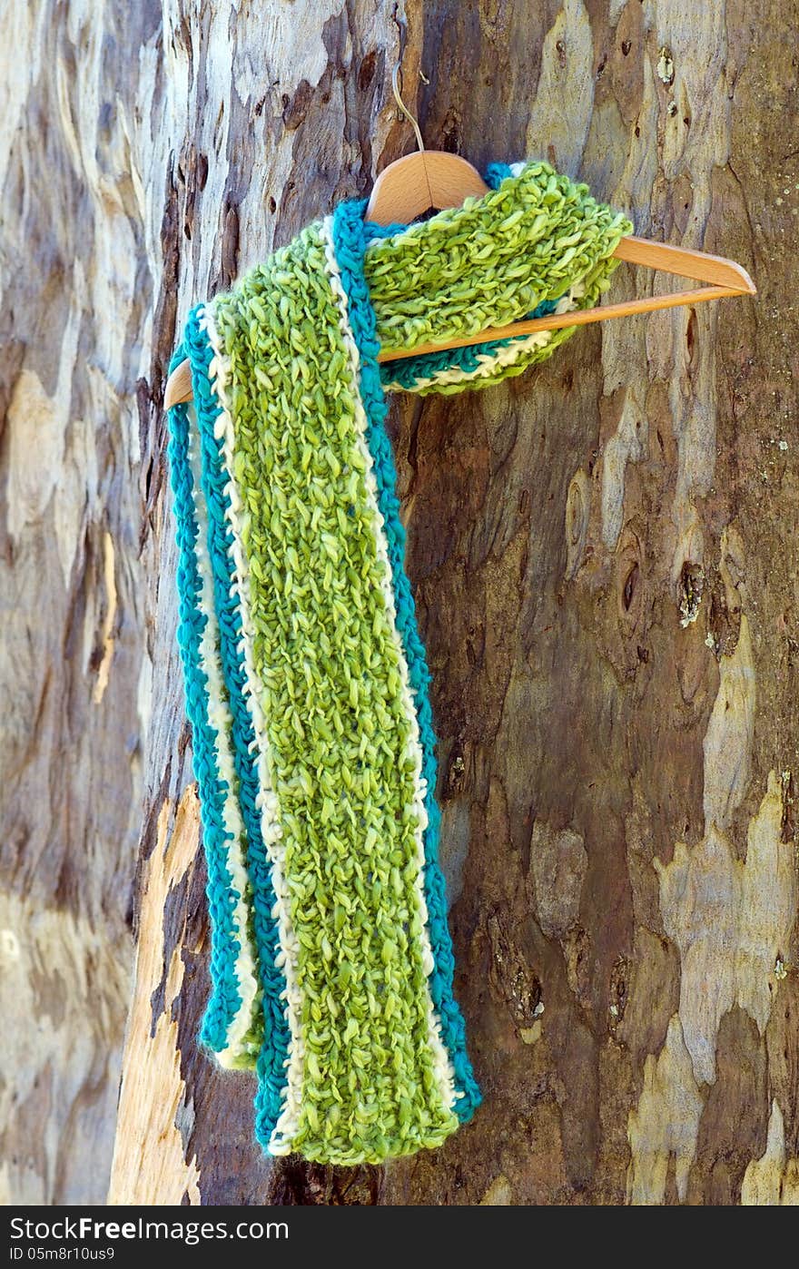 Bright green and blue crochet scarf, fluffy and warm hanging on hanger against wooden tea tree background; concept of winter is coming