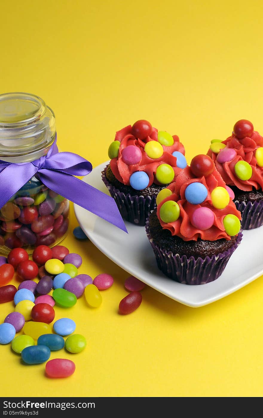 Bright colored candy cupcakes for children birthday, Halloween or Christmas party against a cheerful yellow background, with glass candy jar full of sweets. vertical with copy space for your text here. Bright colored candy cupcakes for children birthday, Halloween or Christmas party against a cheerful yellow background, with glass candy jar full of sweets. vertical with copy space for your text here.