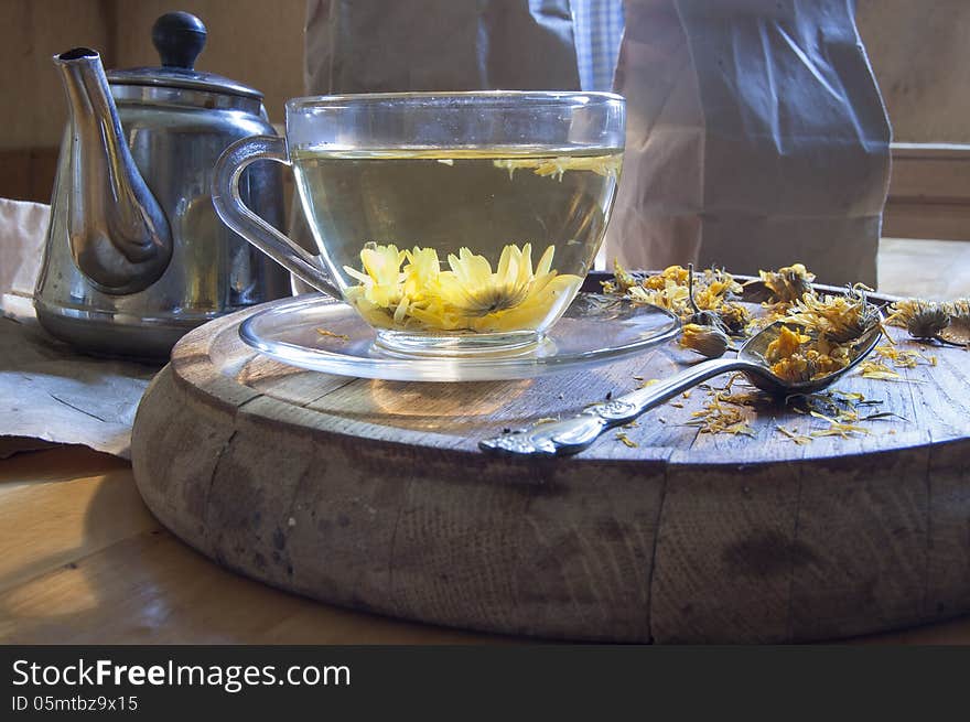 A cup of herbal tea. Near kettle, tea spoon and dried marigold flowers on a wooden board.