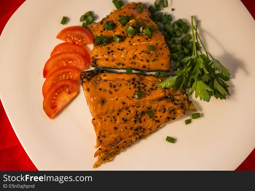 Delicious smoked fish with spices on top served with juicy tomato slices, parsley and green onions. White plate background. Delicious smoked fish with spices on top served with juicy tomato slices, parsley and green onions. White plate background.