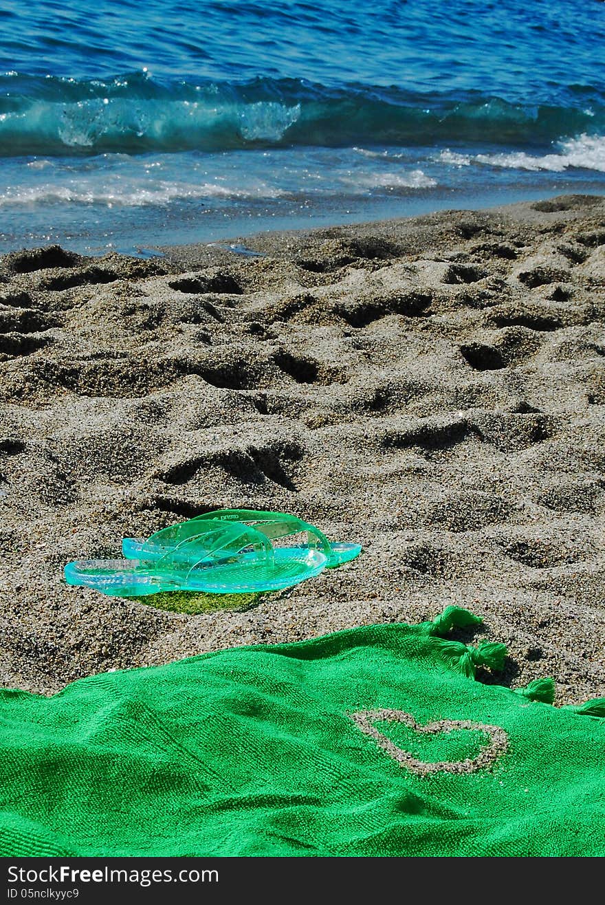 Green flipflop on a beach covered by sand, towel with a heart made by sand and the sea in the background. Green flipflop on a beach covered by sand, towel with a heart made by sand and the sea in the background