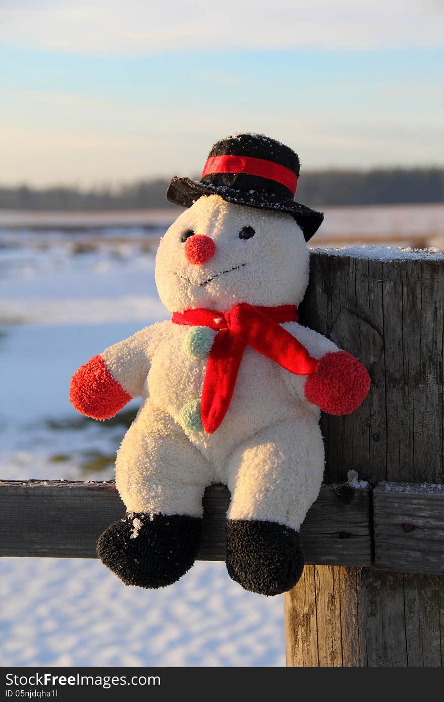 Toy snowman sitting on a wooden fence outdoors at sunset in winter. Toy snowman sitting on a wooden fence outdoors at sunset in winter