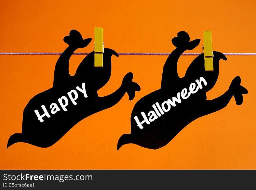 Happy Halloween greeting written across black ghosts cards hanging from pegs on a line against an orange background.