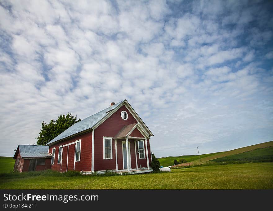 Little red schoolhouse on green grass with blue clouded sky in background.