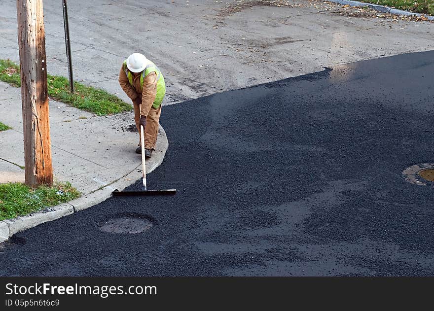 Construction worker putting the finishing touches on repaving a street by manually spreading the asphalt on top.