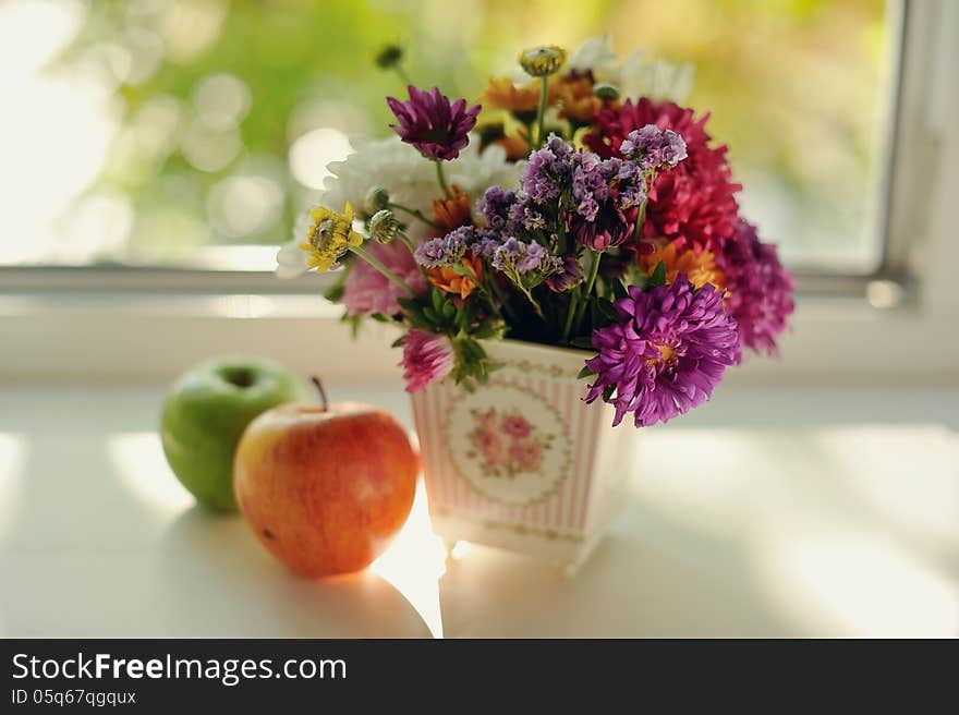 On the window sill is a bouquet of wild flowers in a beautiful decorative vase and some apples are. On the window sill is a bouquet of wild flowers in a beautiful decorative vase and some apples are