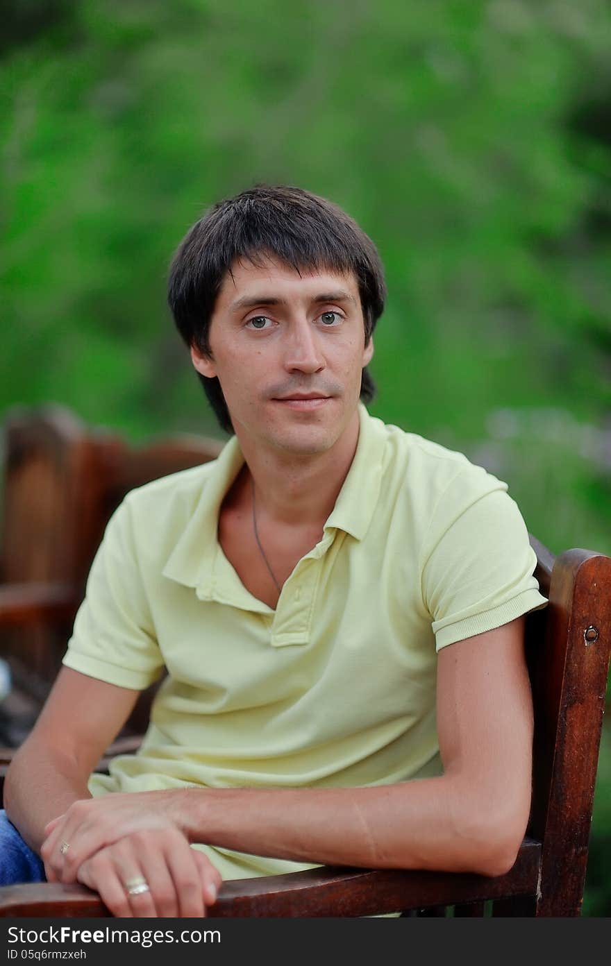 A young man in a yellow shirt is sitting on a wooden chair in nature. A young man in a yellow shirt is sitting on a wooden chair in nature