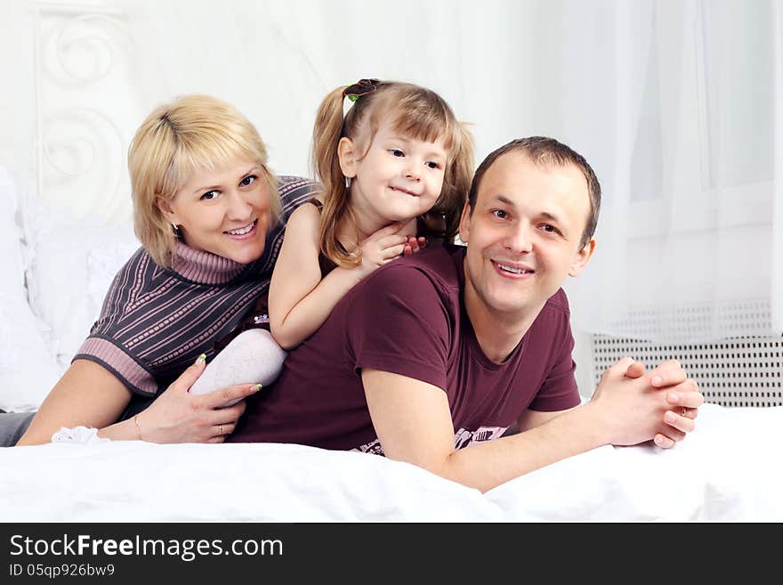 Little girl, father and mother lie on white double bed and smile. Focus on woman. Little girl, father and mother lie on white double bed and smile. Focus on woman.