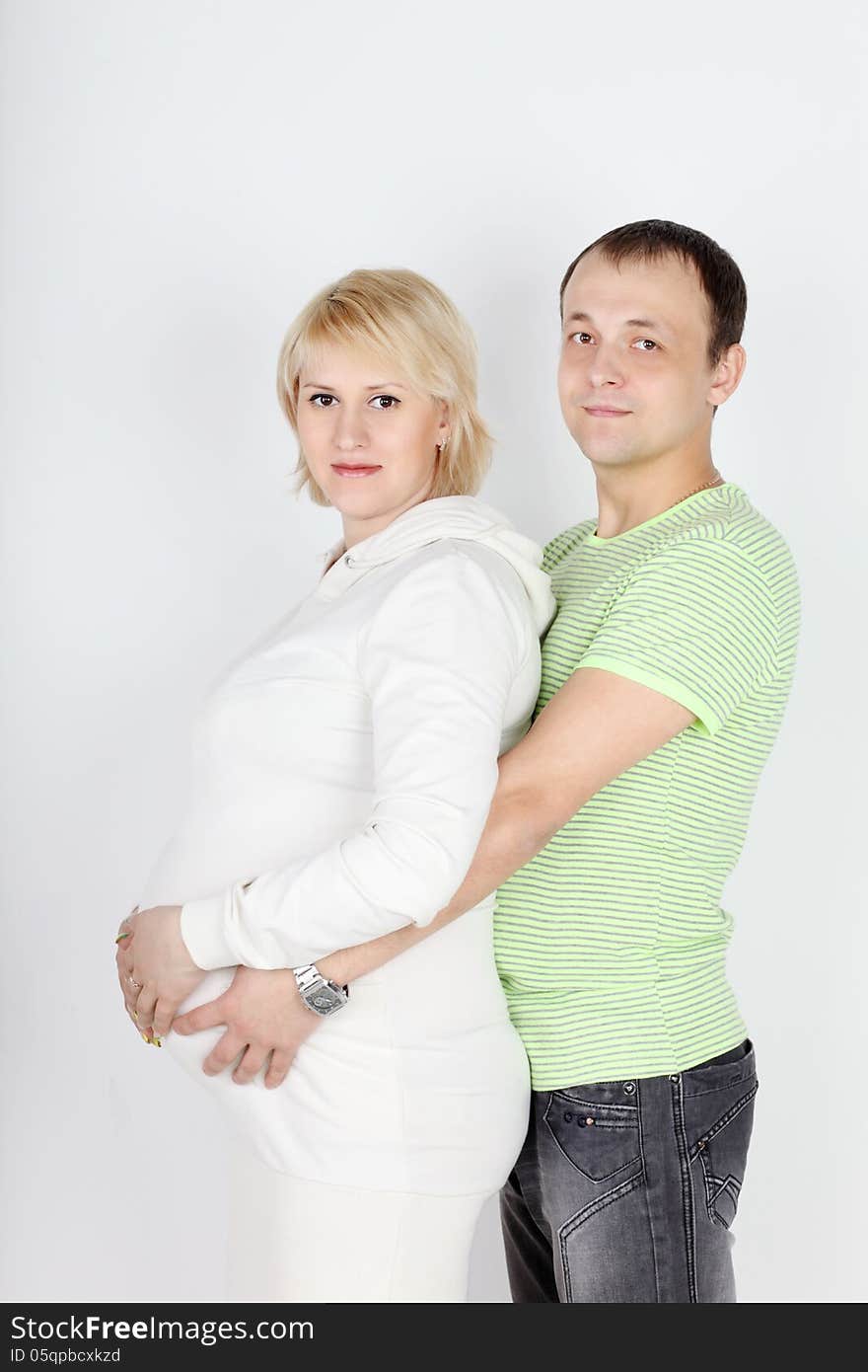 Happy pregnant wife in white and husband hold belly on white background.