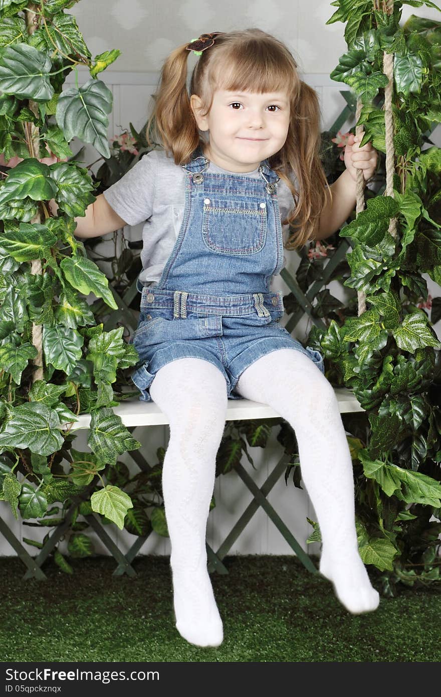 Happy little girl in denim jumpsuit sits on swing