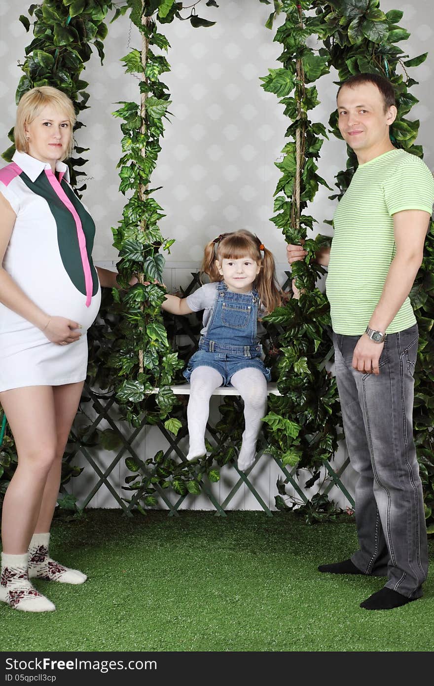 Little girl sits on swing under green ivy and father and pregnant mother swing she. Little girl sits on swing under green ivy and father and pregnant mother swing she.