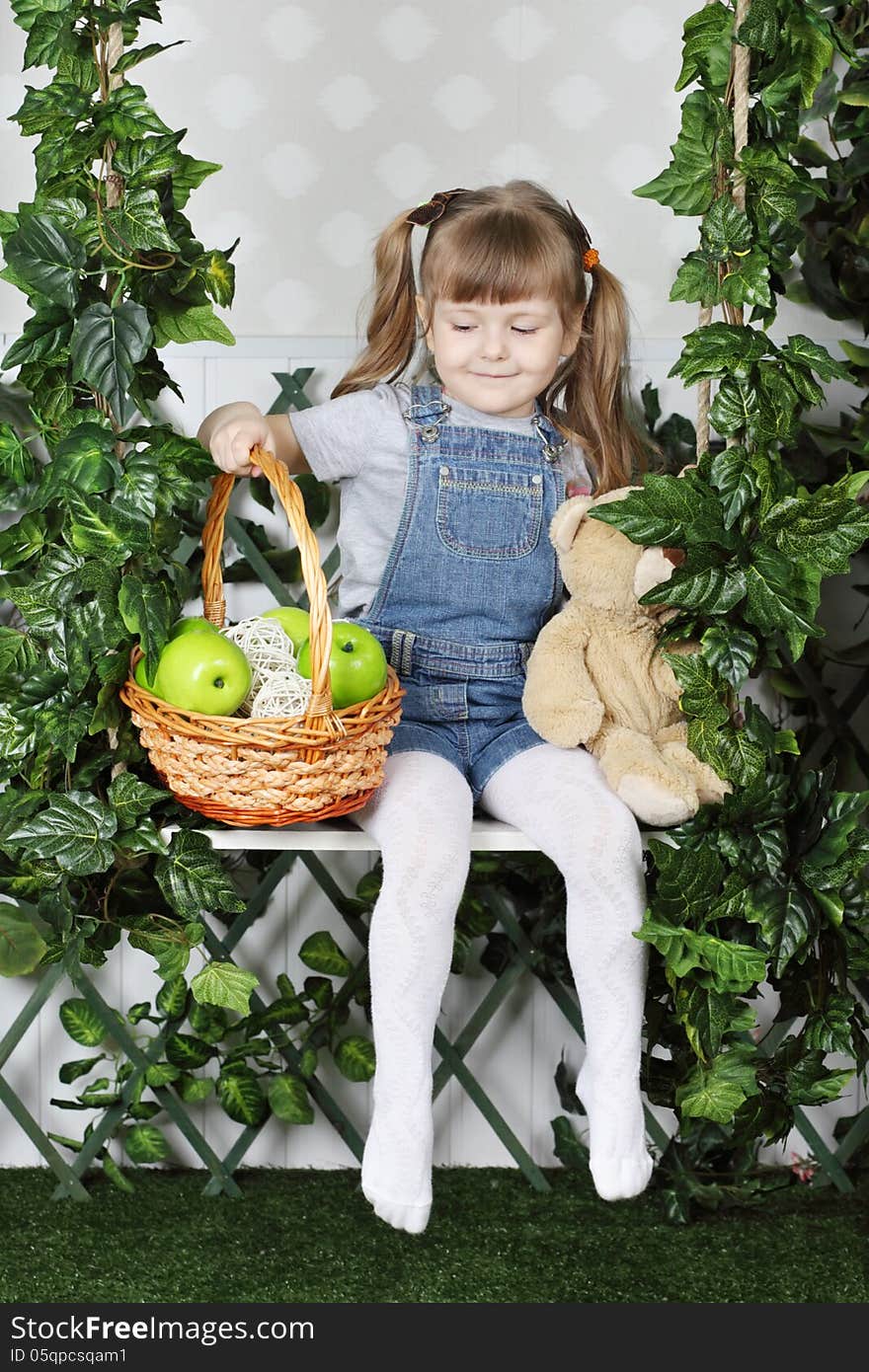 Happy little girl sits on swing under green ivy with teddy bear and basket with apples. Happy little girl sits on swing under green ivy with teddy bear and basket with apples.