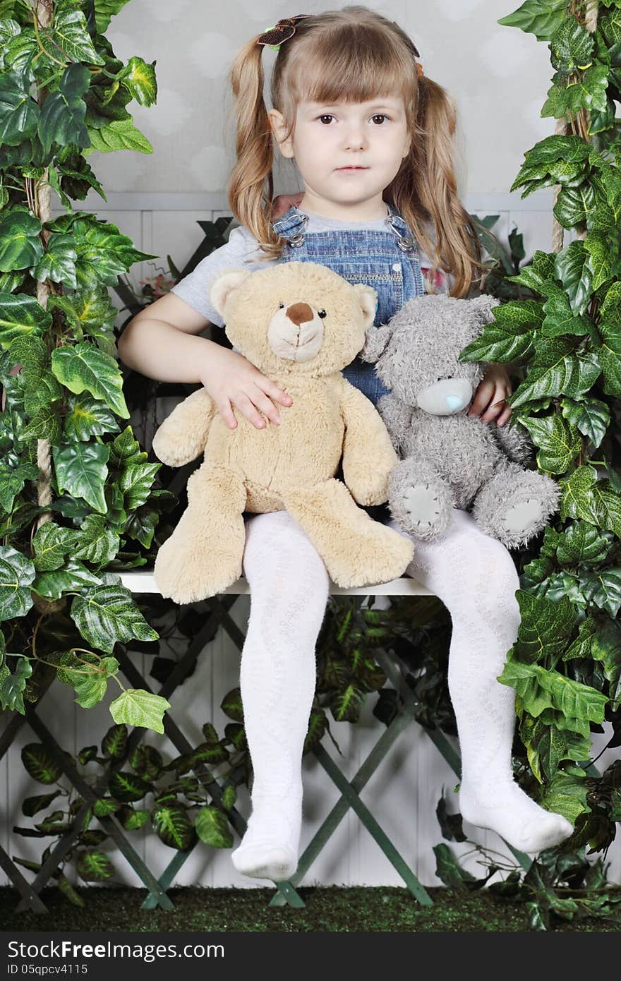 Tired little girl in denim jumpsuit sits on swing under green ivy with two teddy bears.