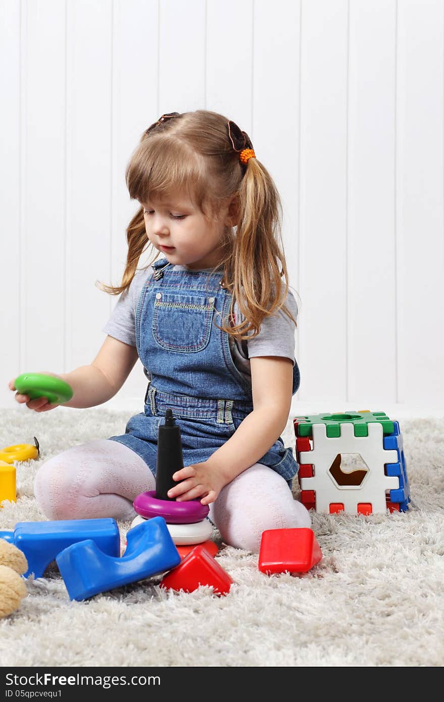 Happy little girl gathers pyramid among multi-colored toys on soft carpet at home. Happy little girl gathers pyramid among multi-colored toys on soft carpet at home.