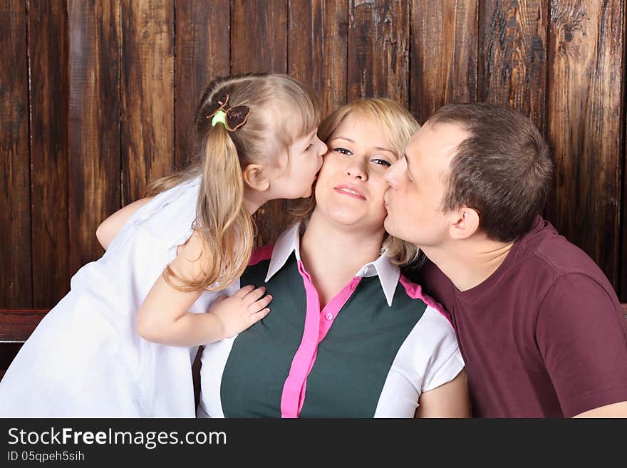 Father and little daughter kiss mother on bench next to wooden wall.