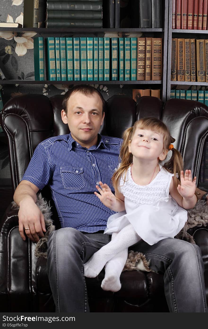 Man and her daughter sit in leather armchair next to shelves with lots of books.