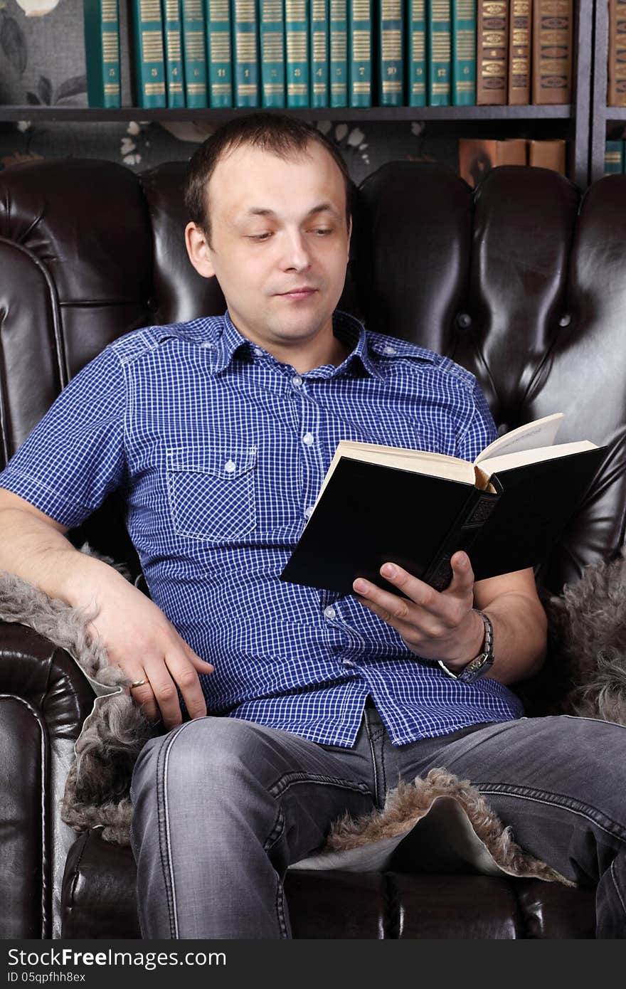 Young man sits in leather armchair and reads book next to shelves with lots of books.