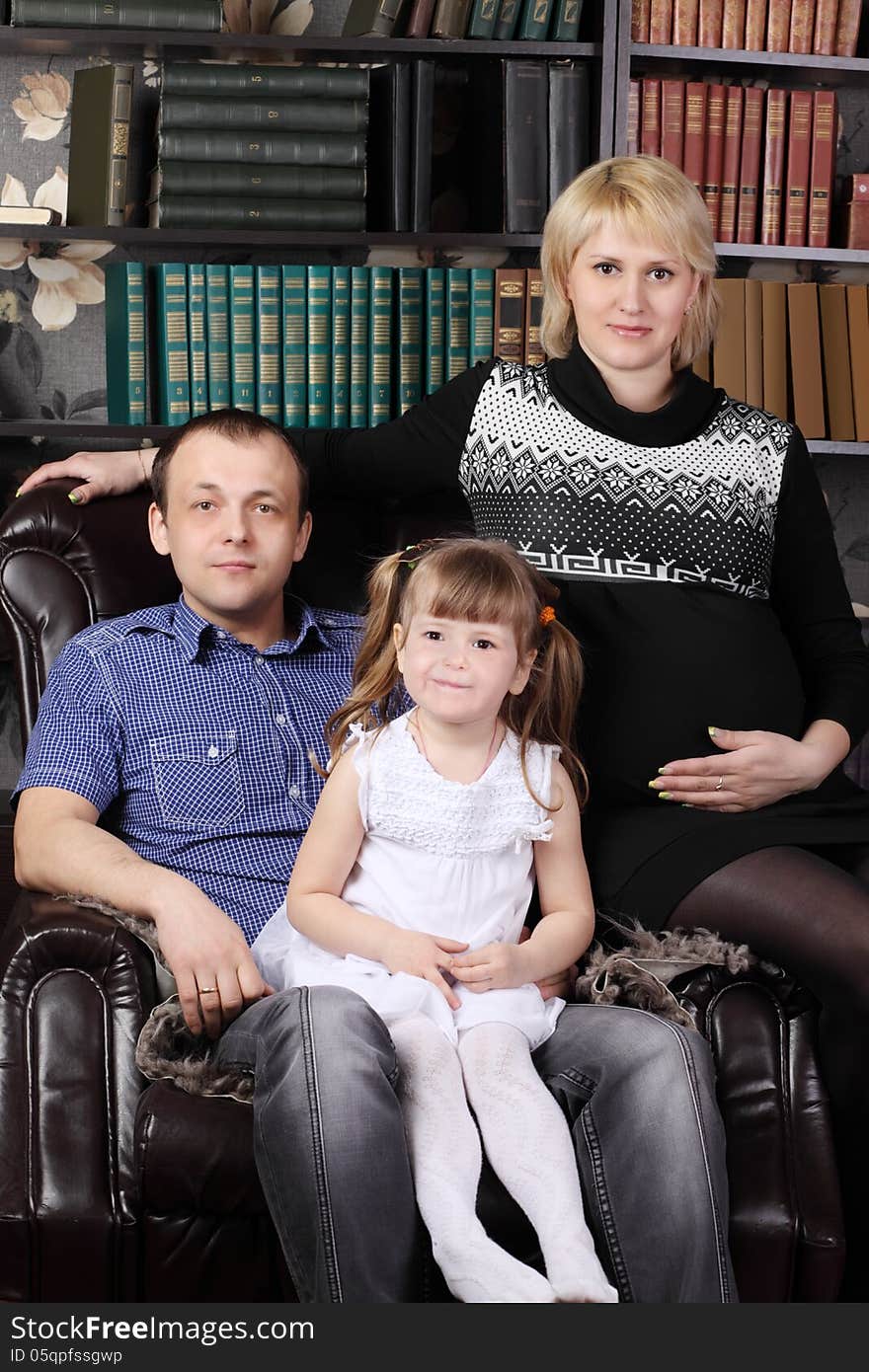 Father, mother and daughter sit in leather armchair next to shelves with books. Father, mother and daughter sit in leather armchair next to shelves with books.