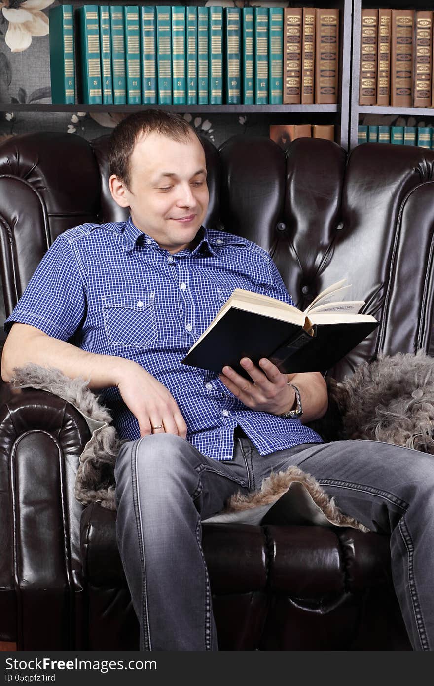 Smiling man sits in leather armchair and reads book next to shelves with lots of books.