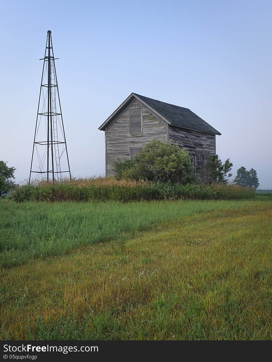 An old farm house and a windmill tower are still standing in a hay field.