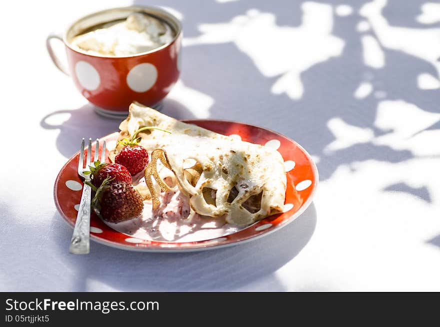 Pancakes with strawberries and sour cream on the white tablecloth. Next to a cup of coffee. All lit bright sun. On the tablecloth visible shadows from tree leaves. Pancakes with strawberries and sour cream on the white tablecloth. Next to a cup of coffee. All lit bright sun. On the tablecloth visible shadows from tree leaves.