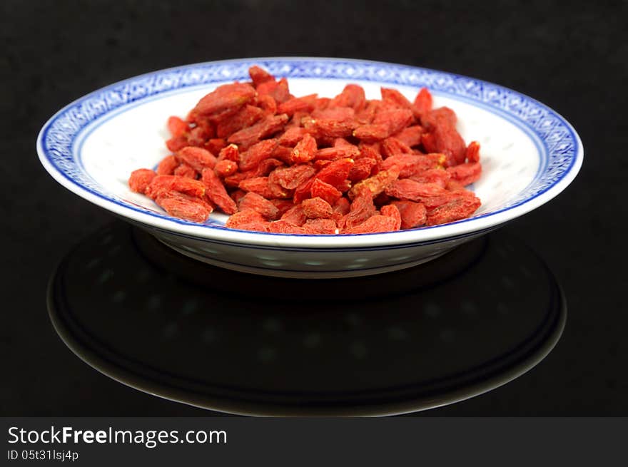The photograph shows the goji berry placed on a porcelain saucer placed on a black mirrored background. The photograph shows the goji berry placed on a porcelain saucer placed on a black mirrored background.