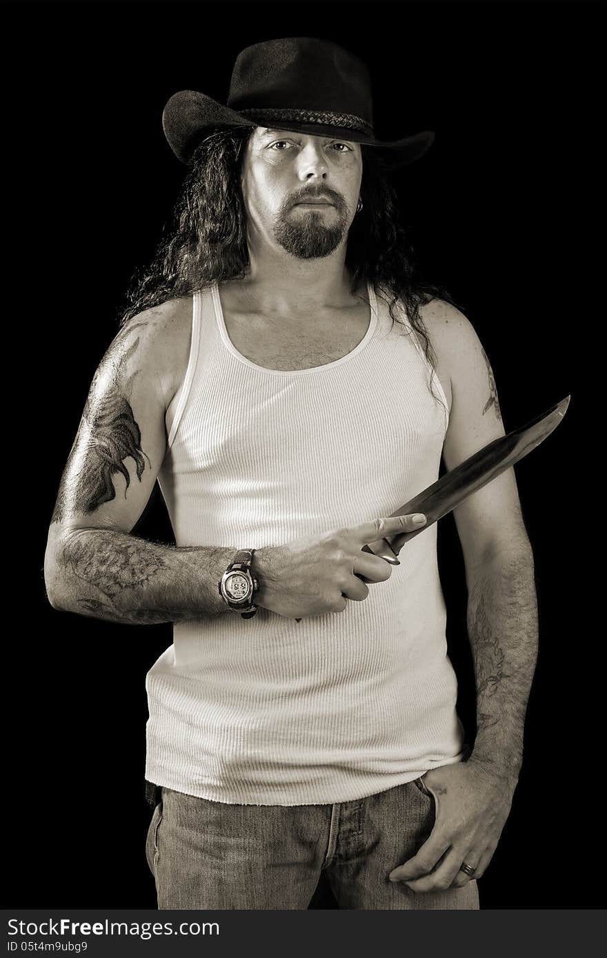 Tough man with long hair wearing a cowboy hat holding a large knife in monochrome isolated on black. Tough man with long hair wearing a cowboy hat holding a large knife in monochrome isolated on black