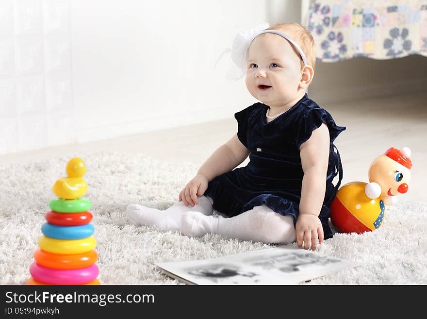 Little cute baby in dress sits on soft carpet among toys and smiles.