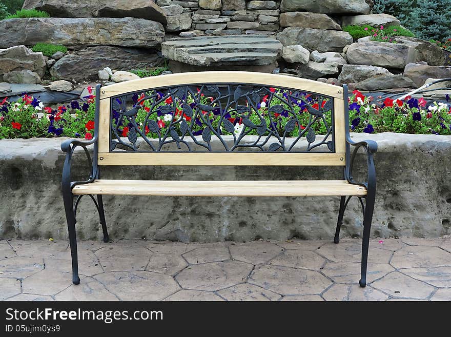 Ornate metal and wood park bench, decorated with birds and leaves, shown with background of stones and colorful flowers. Ornate metal and wood park bench, decorated with birds and leaves, shown with background of stones and colorful flowers.