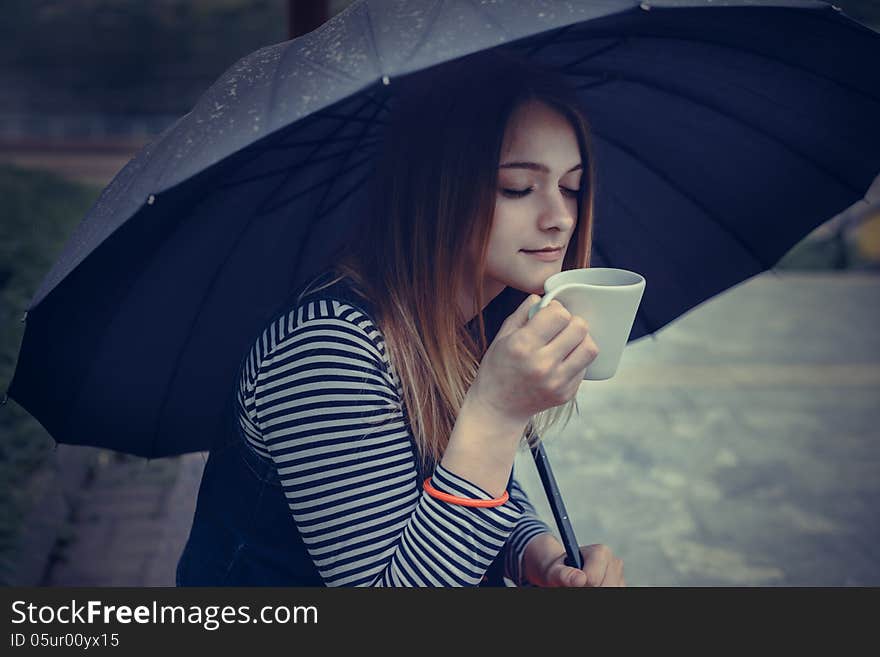 Beautiful girl during a rain under umbrella drinks hot coffee