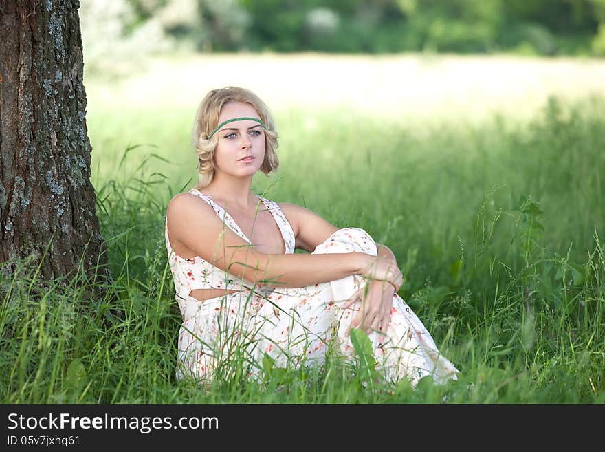 Beautiful girl in a long bright dress sitting in the grass near a tree trunk. Beautiful girl in a long bright dress sitting in the grass near a tree trunk