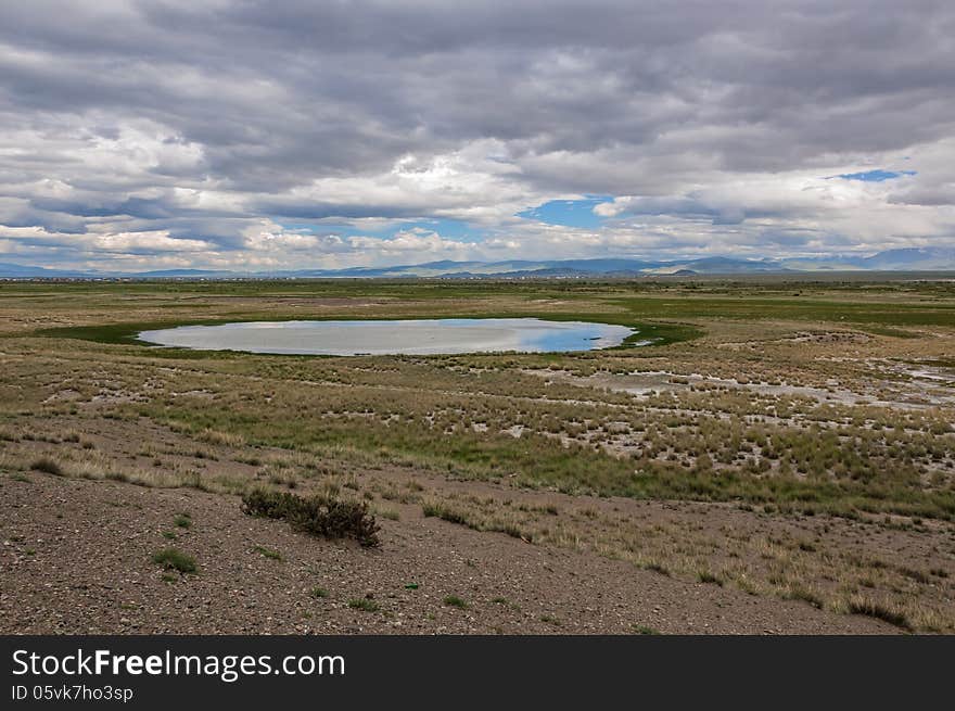 A small lake in the steppe against the backdrop of the mountains, the sky and clouds. In the lake reflects the sky and clouds. A small lake in the steppe against the backdrop of the mountains, the sky and clouds. In the lake reflects the sky and clouds.