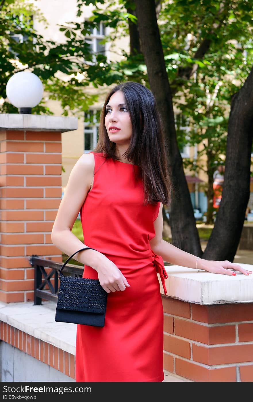 The girl in a red dress stands near a stone fence half-turned