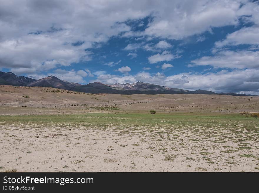 Steppe desert landscape with mountains against the background of blue sky with clouds. Dry land with rare plants as foreground and the mountains, the sky and clouds as a background. Steppe desert landscape with mountains against the background of blue sky with clouds. Dry land with rare plants as foreground and the mountains, the sky and clouds as a background.