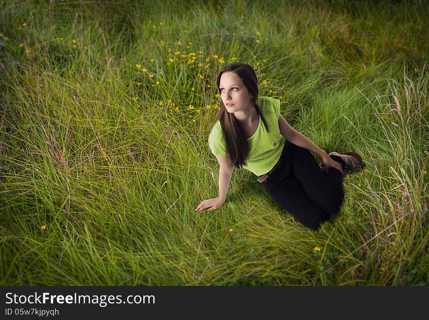 Long hair summer girl wearing green t-shirt and black trousers is sitting in the grass and looking up. Long hair summer girl wearing green t-shirt and black trousers is sitting in the grass and looking up.