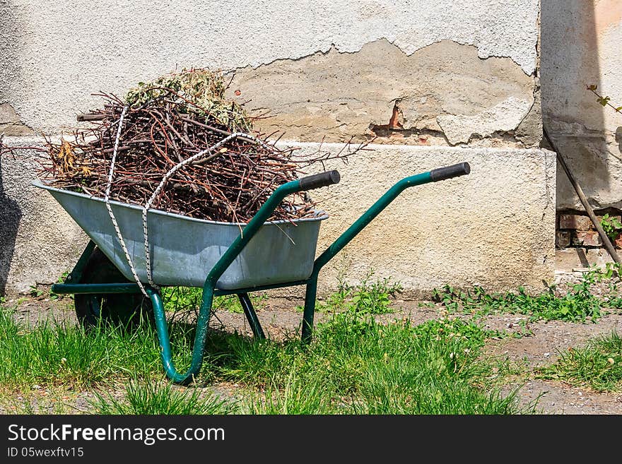 Wheelbarrow filled with cut branches standing in the grass near the old wall. Wheelbarrow filled with cut branches standing in the grass near the old wall