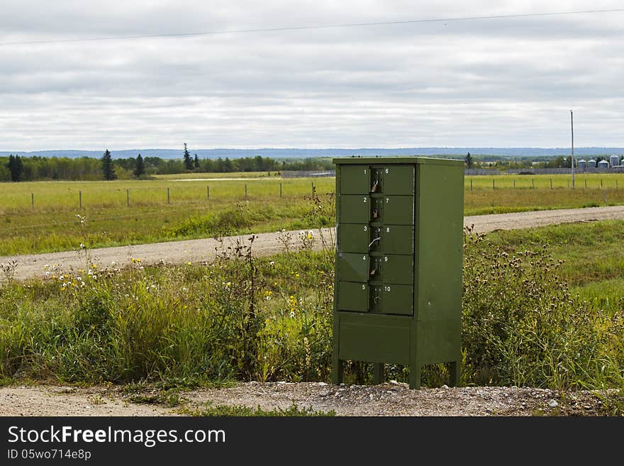 A green metal mailbox with ten boxes beside a road in the countryside with trees and hills in the background. A green metal mailbox with ten boxes beside a road in the countryside with trees and hills in the background