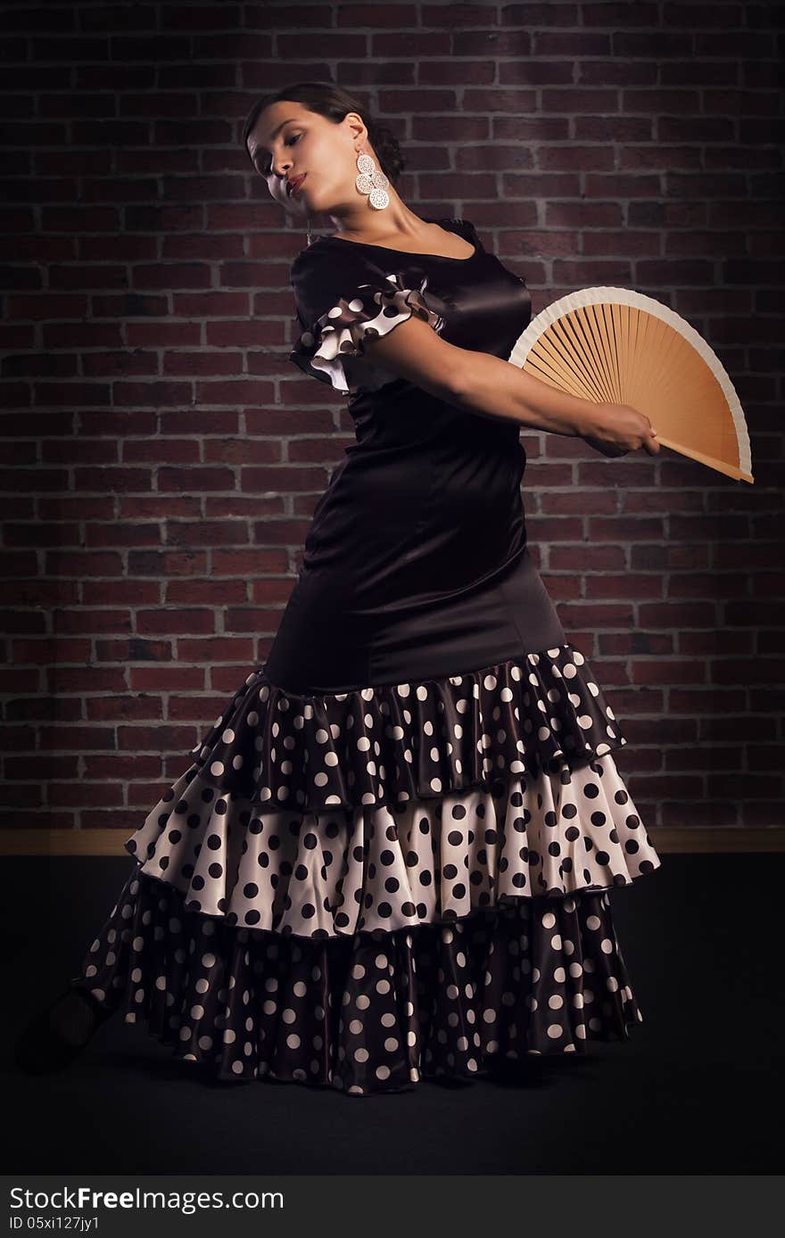 Beautiful young woman wearing brown color silk dress with polka dots is dancing flamenco with spanish hand fan. over red brick wall background. Beautiful young woman wearing brown color silk dress with polka dots is dancing flamenco with spanish hand fan. over red brick wall background.