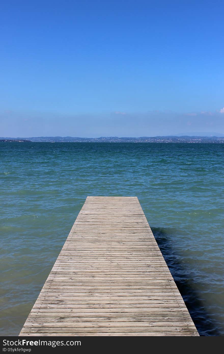 Looking along a wooden jetty out into a lake (Lake Garda in Italy) on a sunny summer day with the other side of the lake visible in the distance.). Looking along a wooden jetty out into a lake (Lake Garda in Italy) on a sunny summer day with the other side of the lake visible in the distance.)