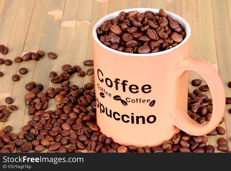 Orange cup with coffee beans is on a wooden table. coffee beans are scattered around the cup. Inscription on the cup coffee, cappuccino. wood background.