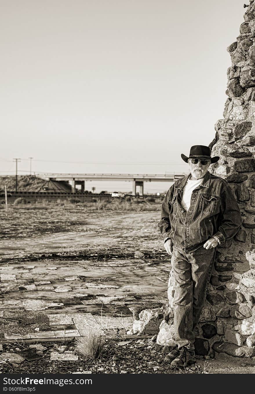 Handsome older man with a gray beard wearing a black cowboy hat leaning against an old stone chimney with a freeway and overpass in the background. Handsome older man with a gray beard wearing a black cowboy hat leaning against an old stone chimney with a freeway and overpass in the background