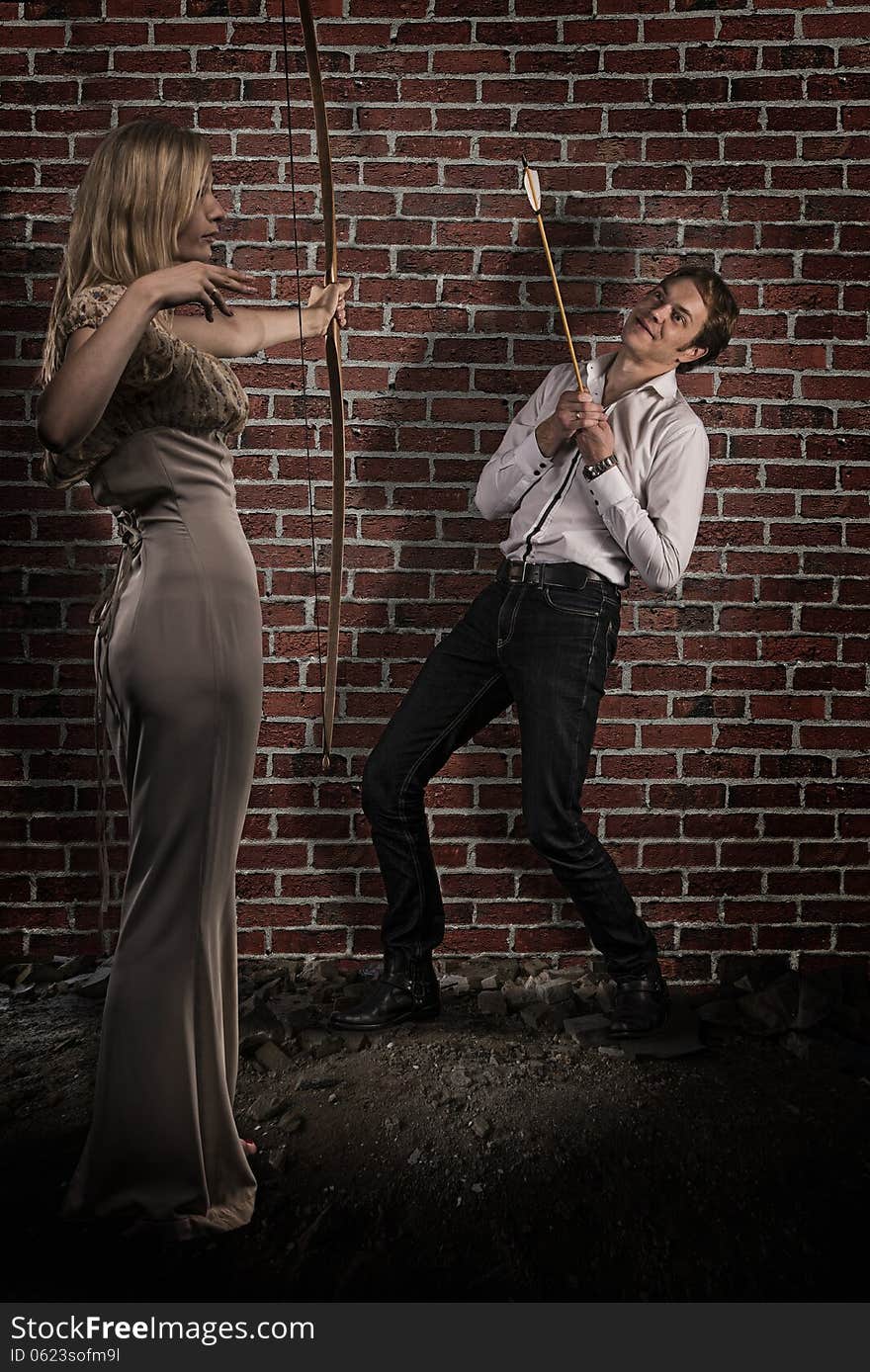 Gorgeous blond hair women wearing elegant dress winning man's heart. Her arrow hits the center of man's heart. Red brick wall background. Gorgeous blond hair women wearing elegant dress winning man's heart. Her arrow hits the center of man's heart. Red brick wall background.