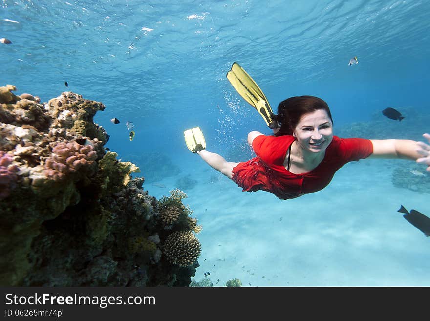 A young woman who is wearing no diving mask but a red dress, swims underwater at a tropical reef and smiles to the camera. A young woman who is wearing no diving mask but a red dress, swims underwater at a tropical reef and smiles to the camera.