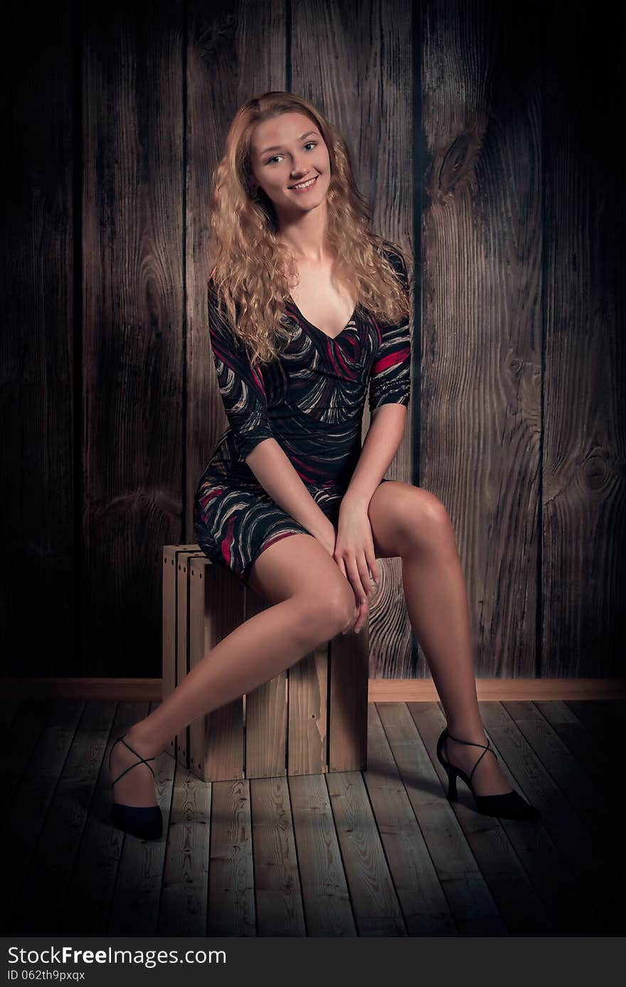 Attractive happy smiling woman with blond long curly hair and fine figure is wearing modern short dress, high-heel shoes and is sitting in a pose on wooden box. Over wooden wall background. Attractive happy smiling woman with blond long curly hair and fine figure is wearing modern short dress, high-heel shoes and is sitting in a pose on wooden box. Over wooden wall background.