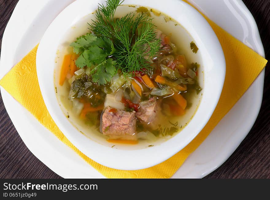 Tasty Homemade Vegetable Soup with Boiled Beef and Greens in White Bowl closeup on White Plate with Yellow Napkin. Top View