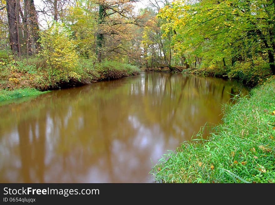 The photograph shows a small river flowing through the forest. It is autumn, the leaves are green, yellow and brown. The photograph shows a small river flowing through the forest. It is autumn, the leaves are green, yellow and brown.